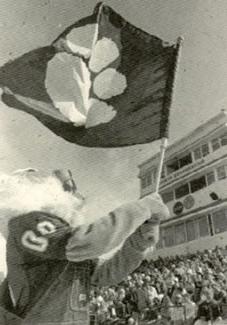 Bobby waves a Bearcat flag during a Football game.  Bobby wears overalls over his classic 00 jersey.